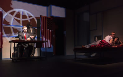 Brutus sits at a table, Cassius stands behnd his left shoulder, ehind oth a screen shows a line-graphic of the globe, an American flag is daped on the open door next to Cassius, and spotlighted to the right of the stage is a fully-clothed Antony lying in bed with Cleopatra, who is under red satin sheets